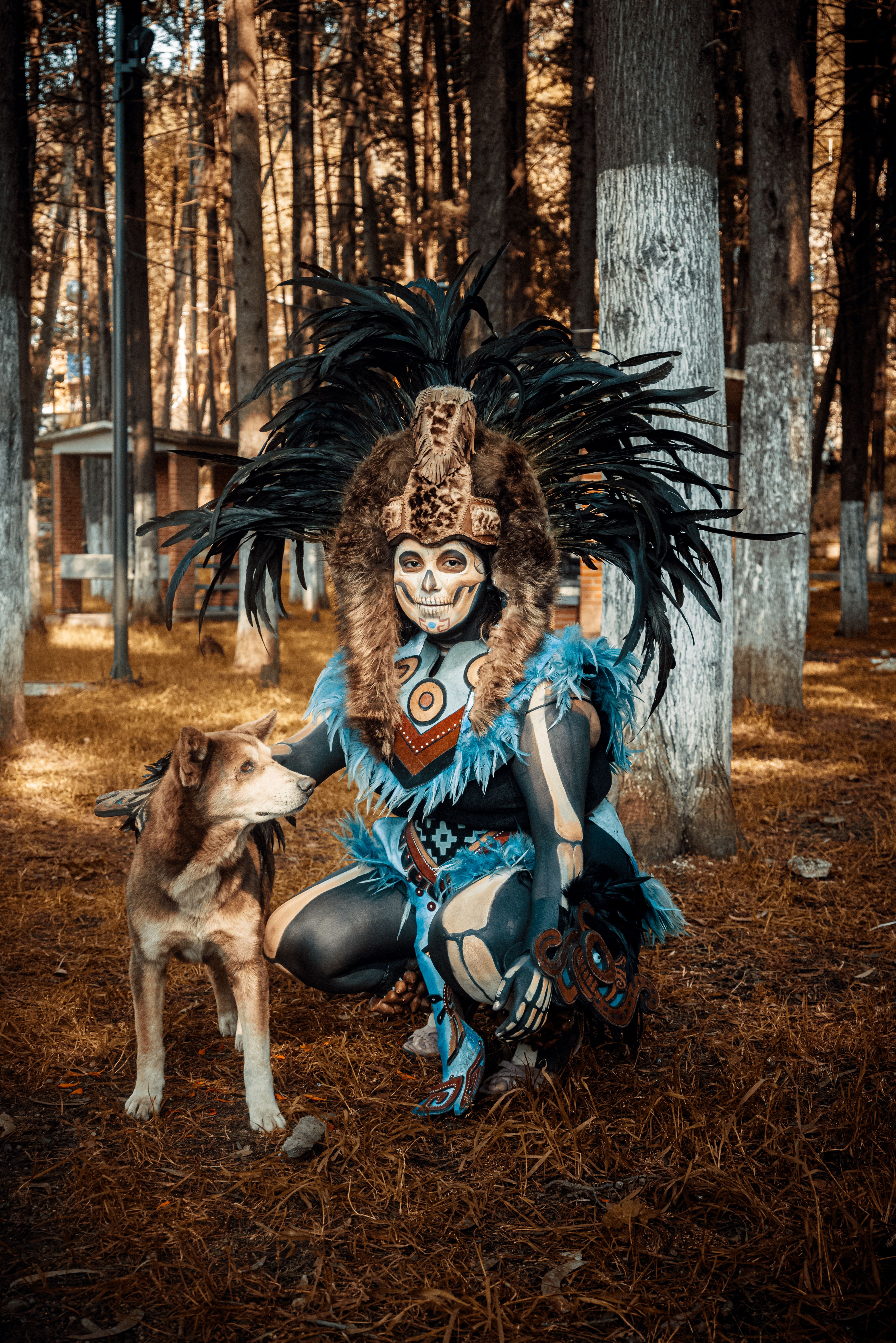 Person in tribal costume and creative makeup with brown dog in a forest setting.