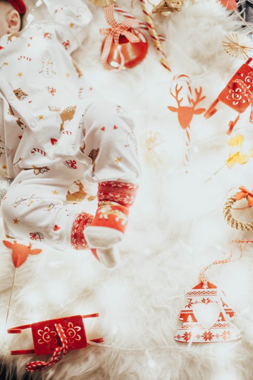 Free Adorable baby in holiday-themed onesie surrounded by warm Christmas decorations and fairy lights. Stock Photo