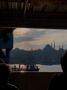 Silhouetted view of the Istanbul skyline featuring mosques at sunset with people and boats on the waterfront.