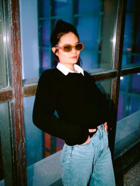 Asian woman in denim and black sweater leaning against glass windows in Montréal.