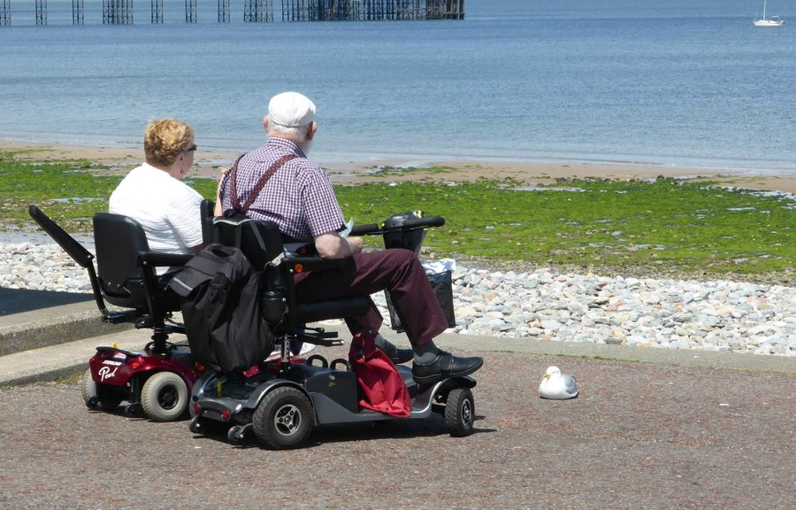 Two people riding mobility scooters together along a seaside path