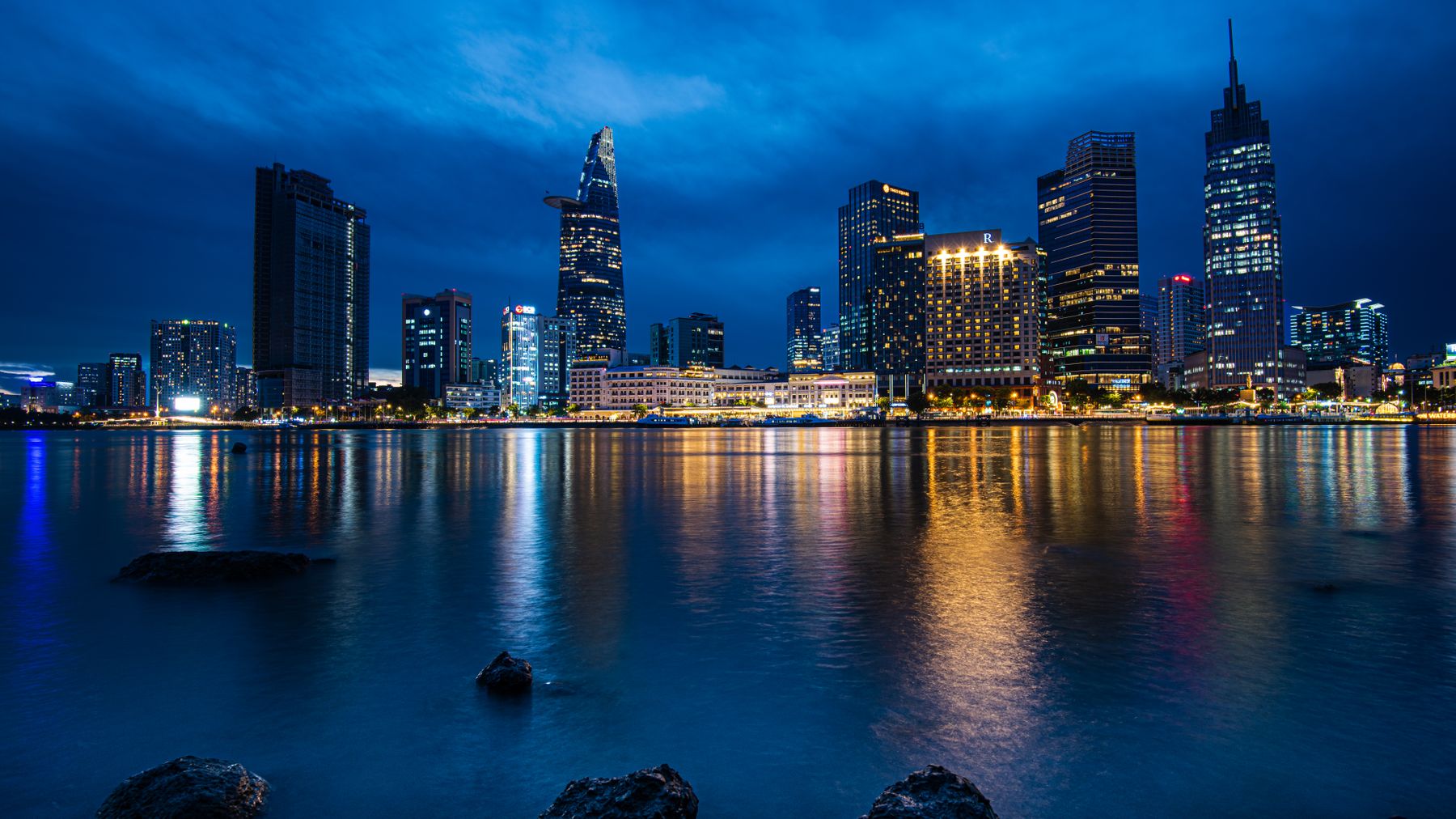 Cityscape of Ho Chi Minh City with skyscrapers and Saigon River at night, reflecting vibrant city lights.