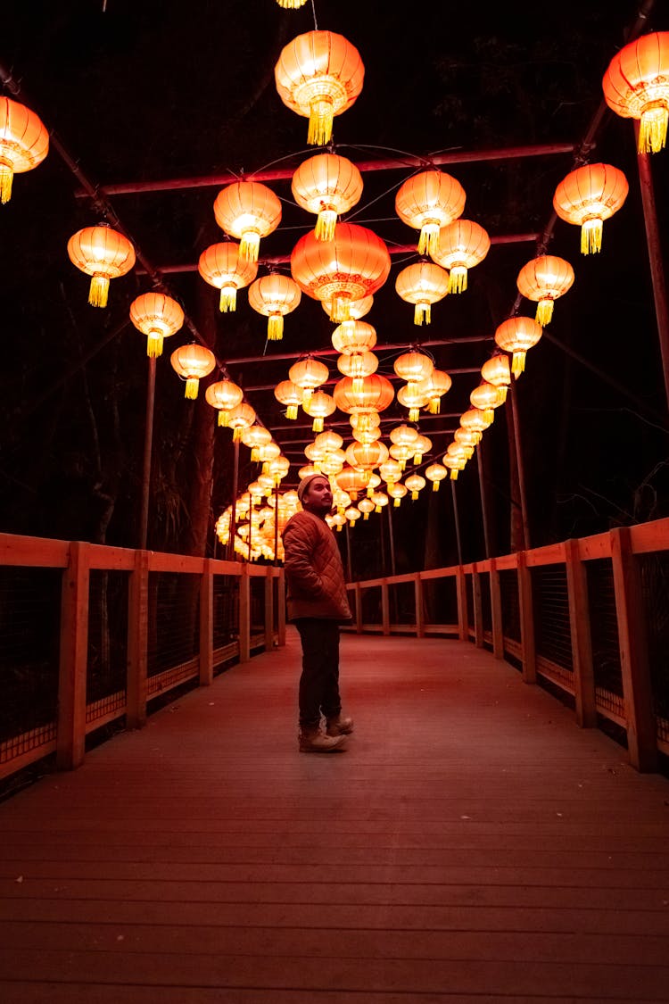 Man In Red Winter Jacket Standing On A Wooden Footbridge With Hanging Chinese Lanterns