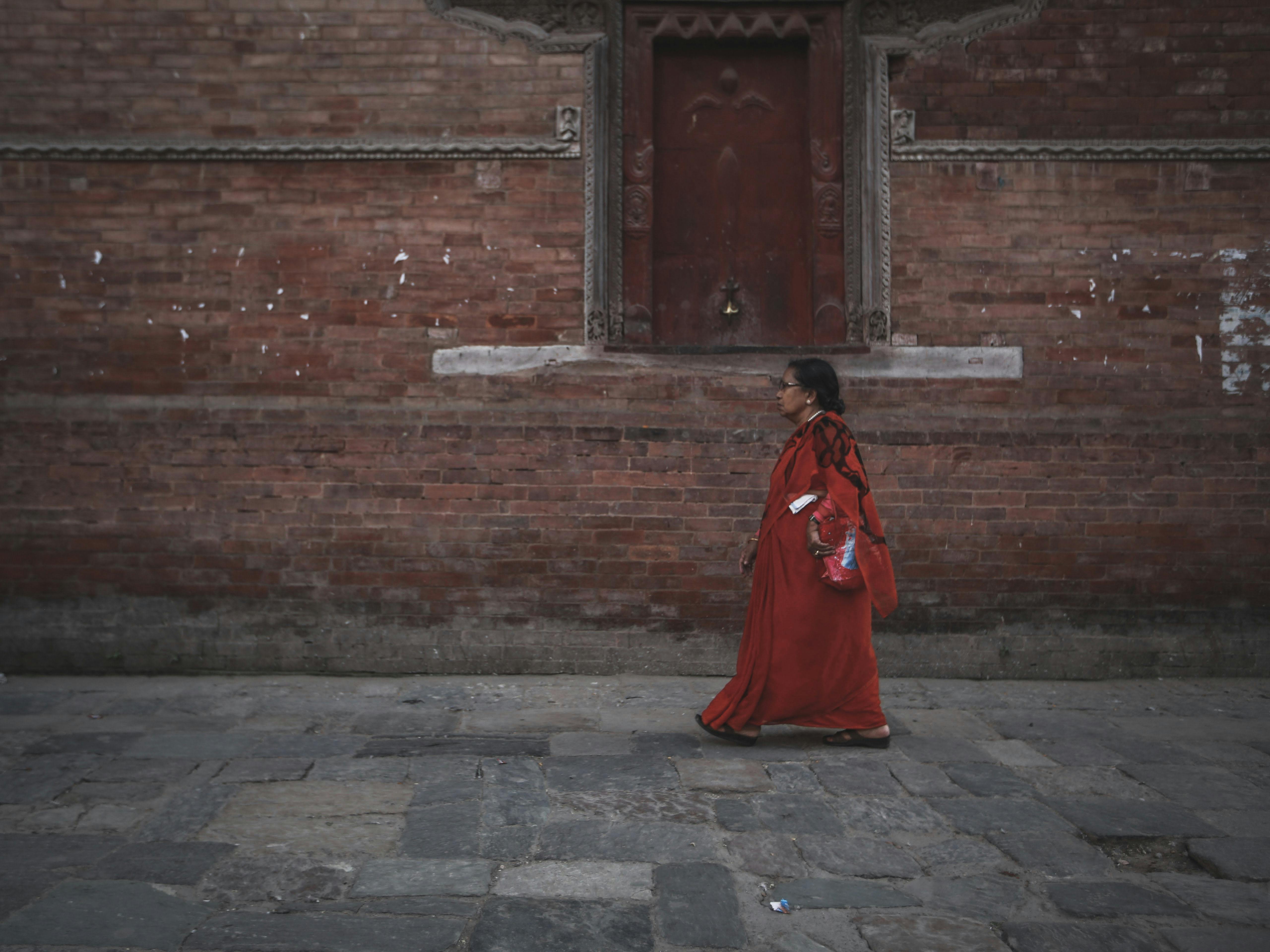 A woman in traditional attire walks alongside a historic brick wall in a quiet street setting.