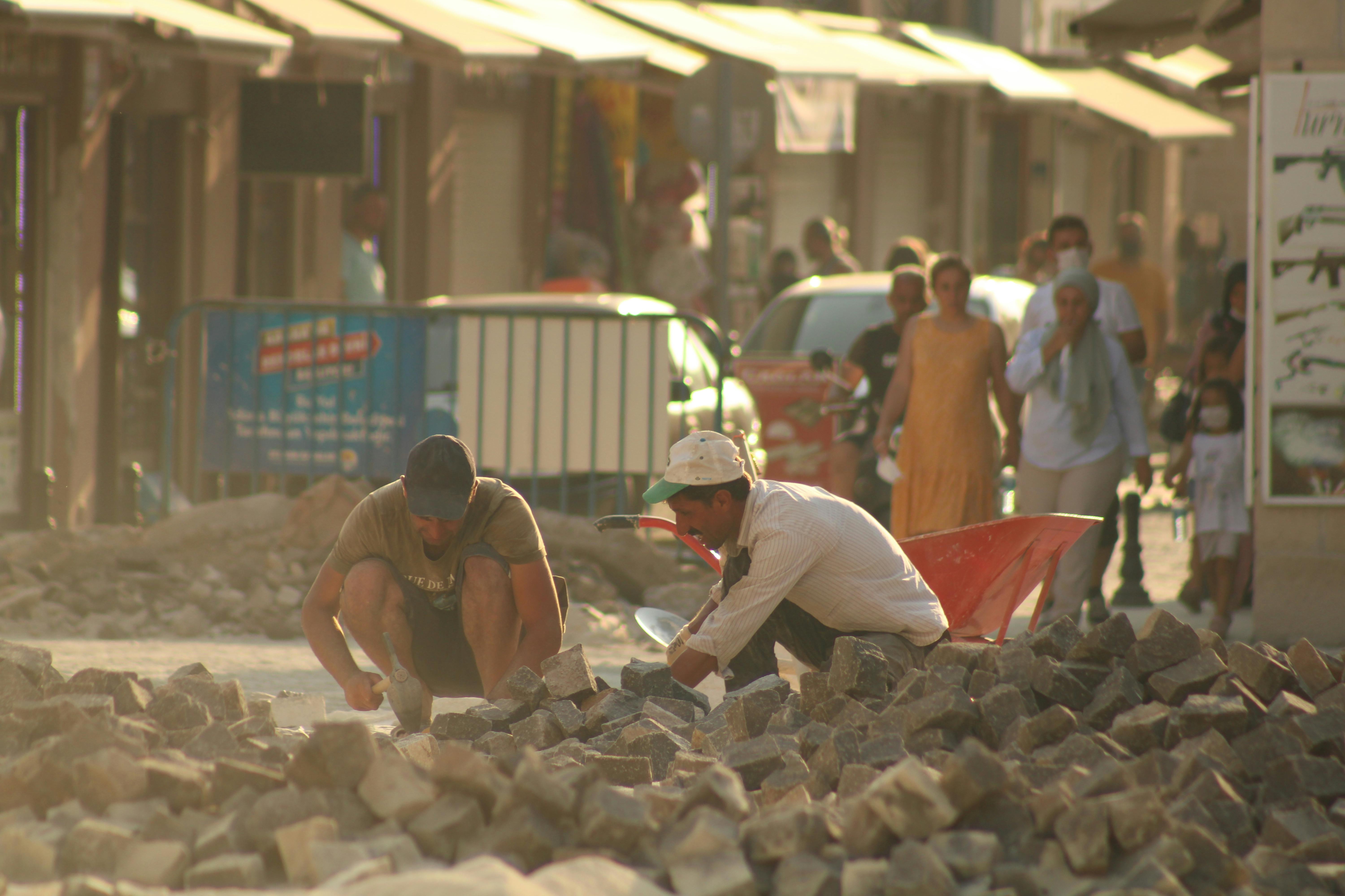 Two workers labor on paving stones in Adana, Turkey, surrounded by urban life