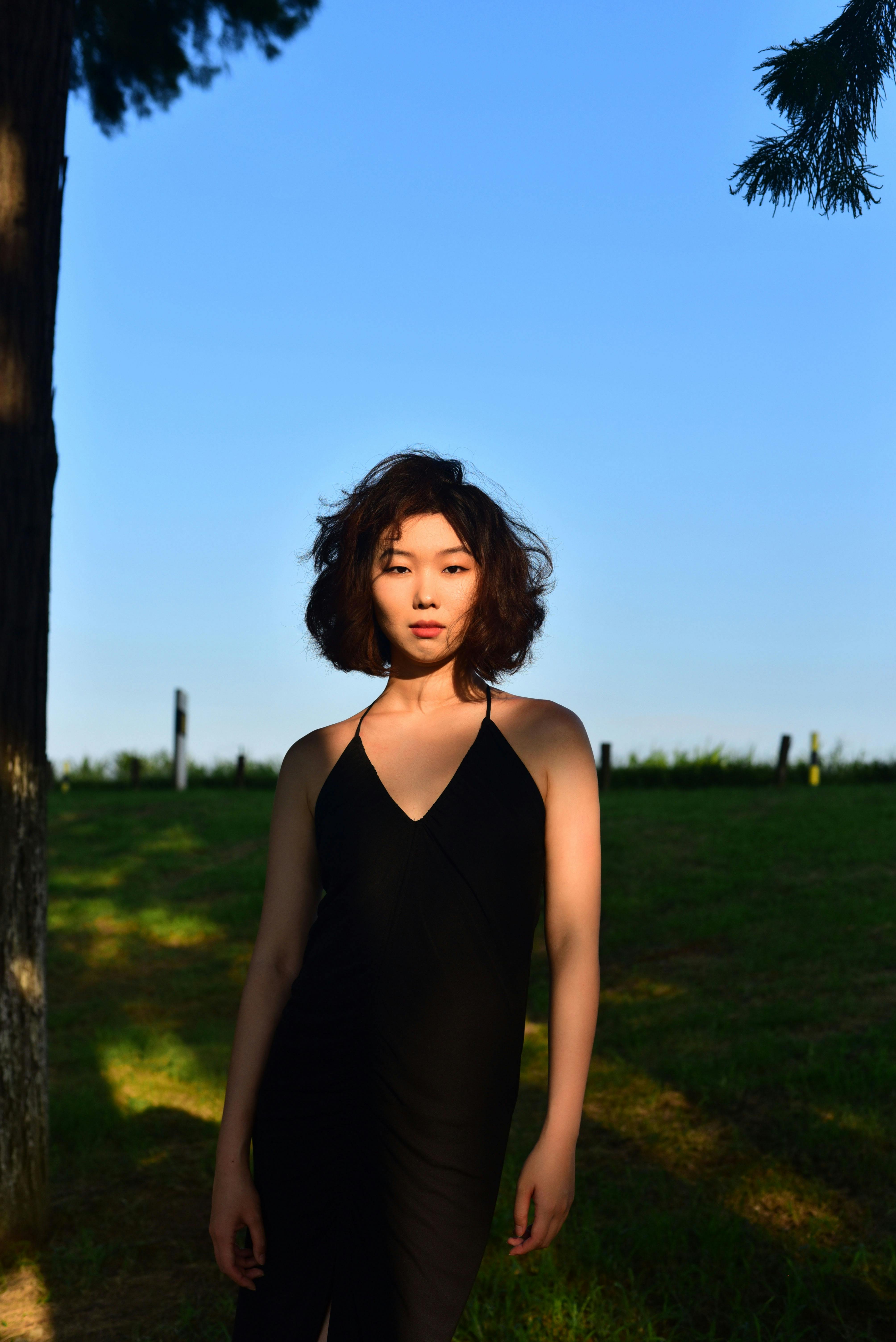 Striking portrait of an Asian woman in a black dress standing outdoors in a sunlit setting.