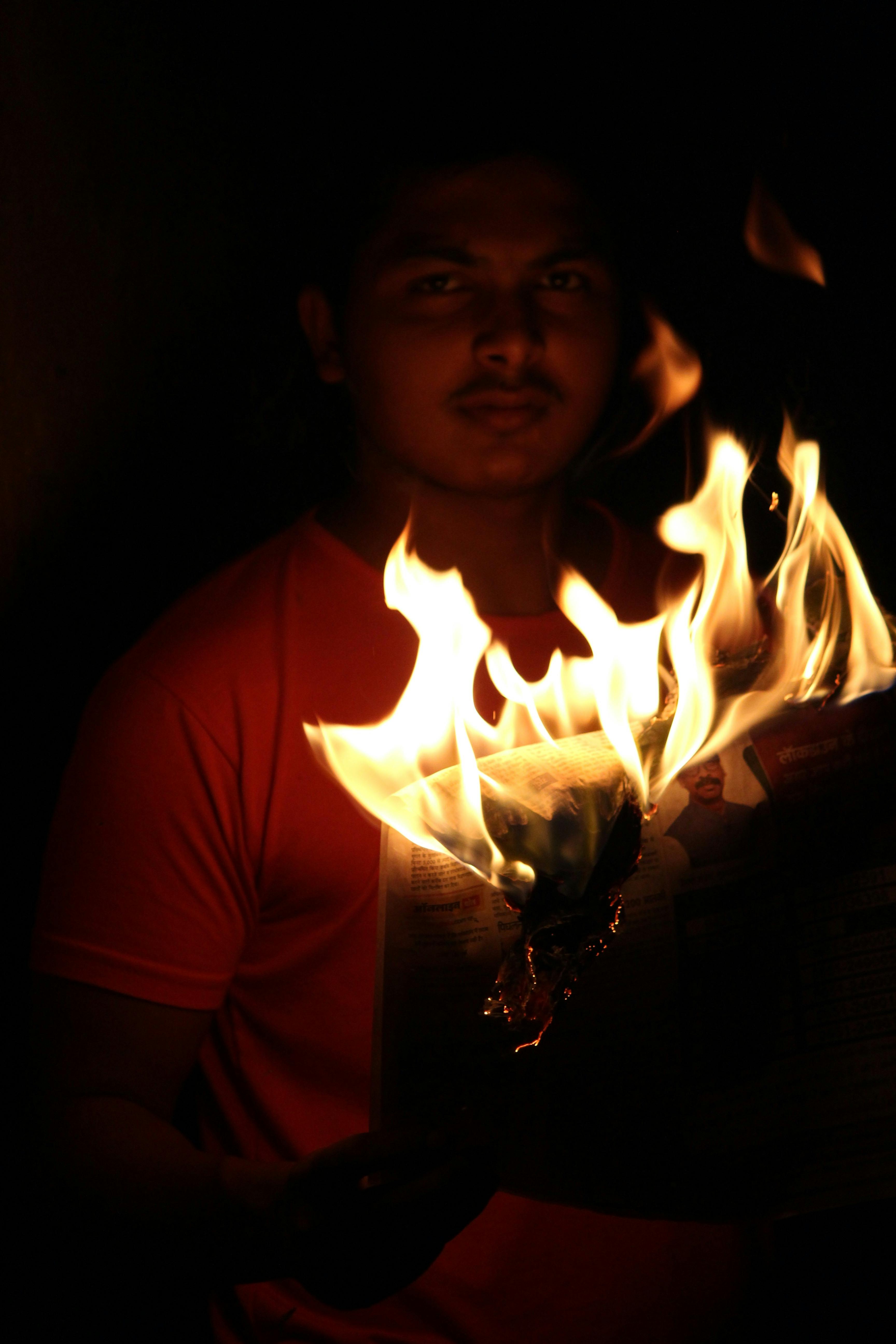 A dramatic shot of a man holding a burning newspaper in the dark, casting warm flames.