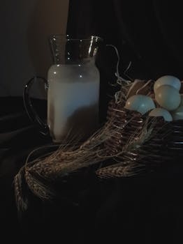 Moody still life featuring a milk jug and eggs in a basket against a dark backdrop.