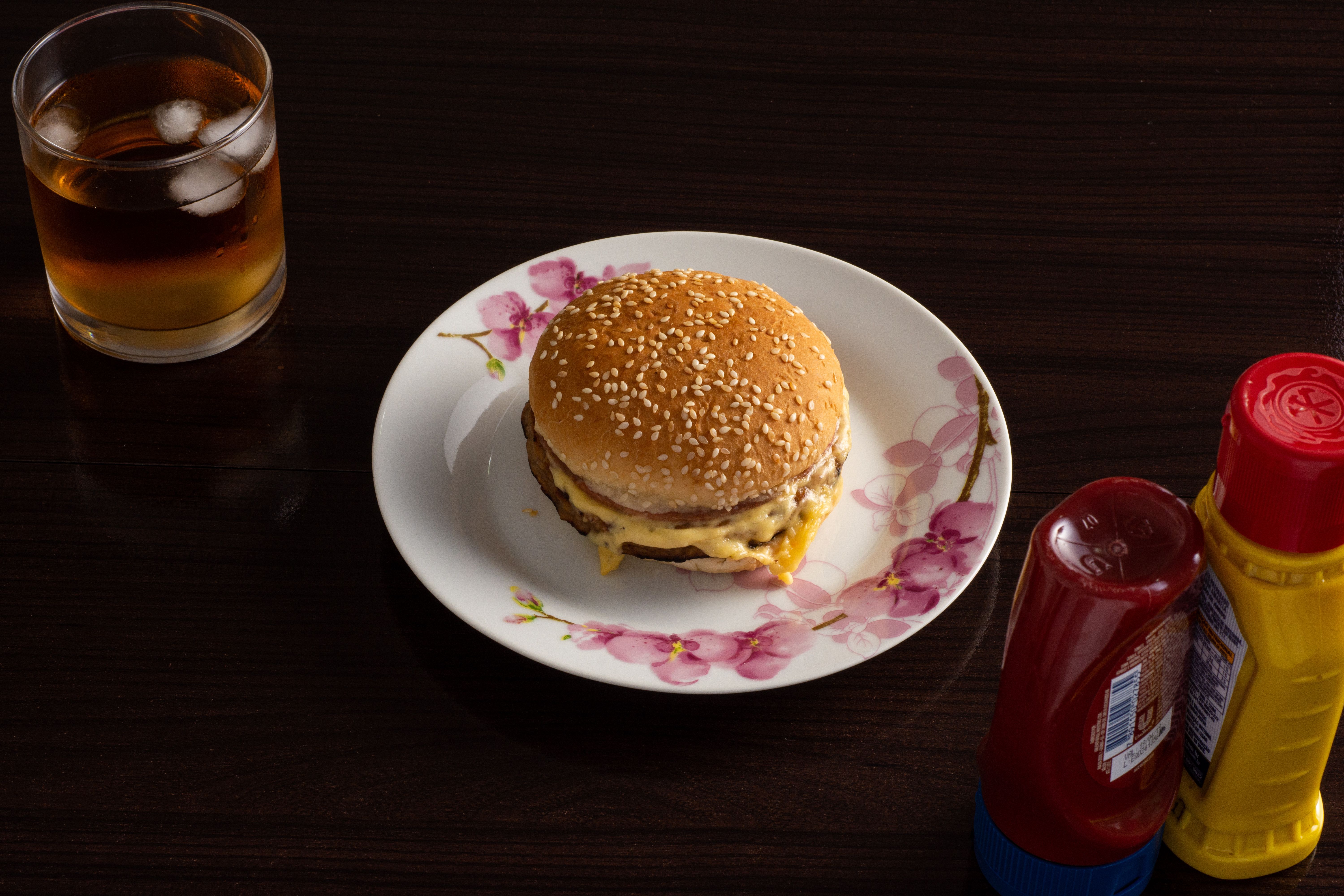 A cheeseburger with condiments and a drink on a dark wooden table.