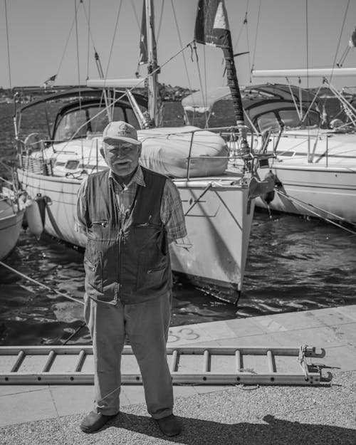 Grayscale Photo of Elderly Man Standing near Sailboats