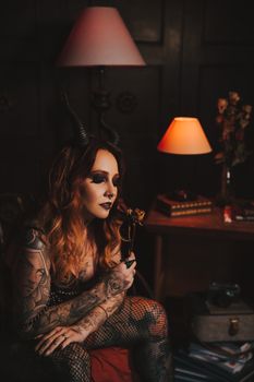 A tattooed woman in gothic attire sits thoughtfully in a dimly lit room.