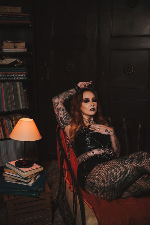 Free A glamorous tattooed woman posing elegantly in a dimly lit room with books and a lamp. Stock Photo