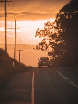 A car travels down a road surrounded by trees and power lines during a vibrant sunset.