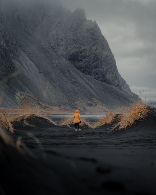 Free A person in a yellow jacket stands on black sand at the base of a towering mountainside. Stock Photo