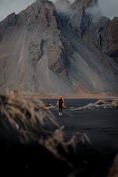 A solitary figure in a yellow jacket hikes on dark terrain with rugged mountains in the background.