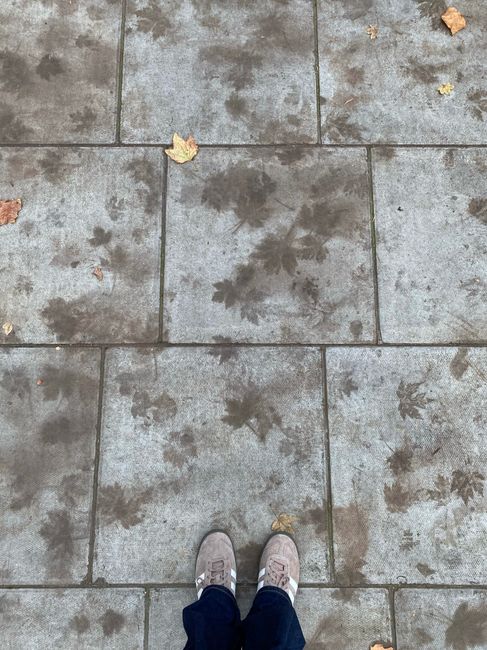 A worn sidewalk with autumn leaf imprints and shoes visible in Greater London.
