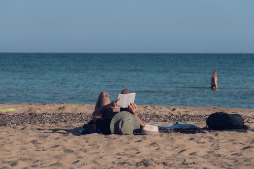 Free Person enjoying a peaceful day reading on a sandy beach with a clear ocean view. Stock Photo