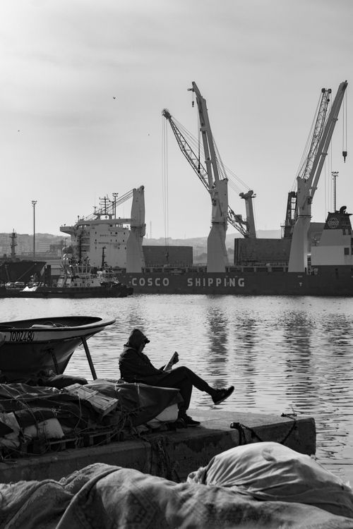 Free A monochrome view of a person relaxing at the harbor with a cargo ship in the background. Stock Photo