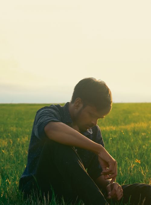 Free A man sitting thoughtfully in a sunny grassy field, evoking solitude and introspection. Stock Photo