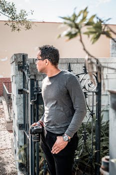 Man in grey shirt holding camera by wrought iron fence in sunny urban setting.