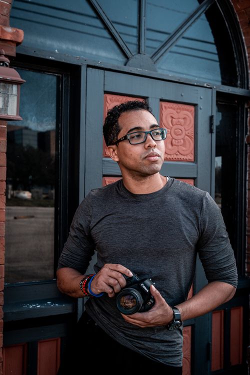 Free Man in gray long sleeve shirt holding a DSLR camera, standing by a decorative door, outdoors. Stock Photo
