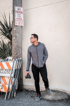 Man in casual attire posing with camera near 'No Parking' sign in city.
