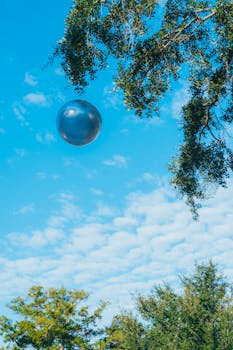 A silver balloon floats amidst a vibrant blue sky and lush green trees, capturing a tranquil outdoor moment.