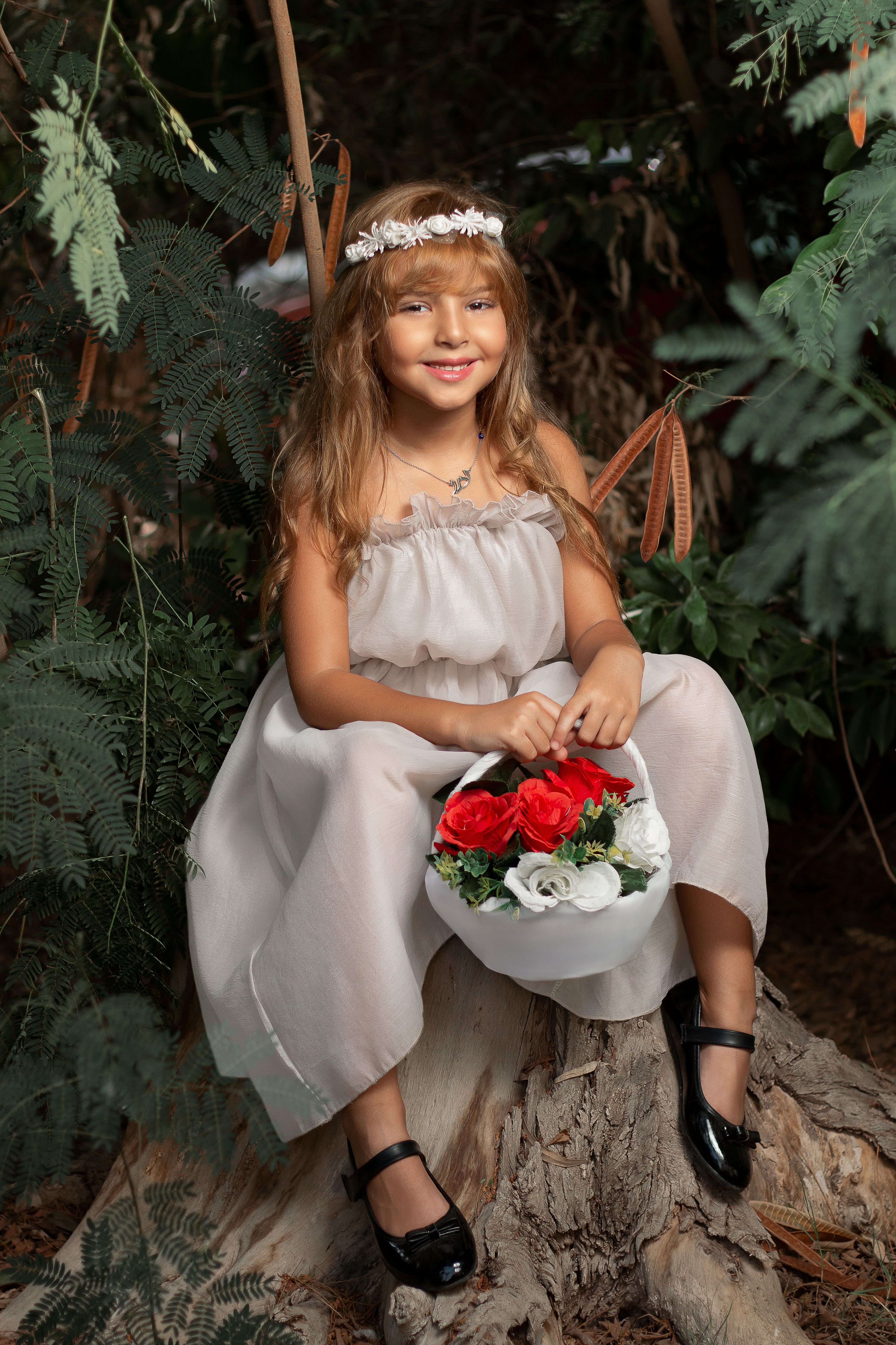 Smiling girl in a white dress with a flower basket, sitting on a tree stump in the woods.