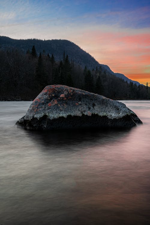 View of a Rock in a Body of Water and Mountains and Trees in the Background at Sunset 
