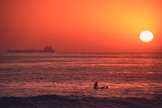 A lone surfer sits in the ocean as the sun sets, creating a vivid orange seascape with an industrial ship on the horizon.
