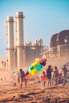 Colorful beach scene juxtaposed against an industrial refinery in the background.