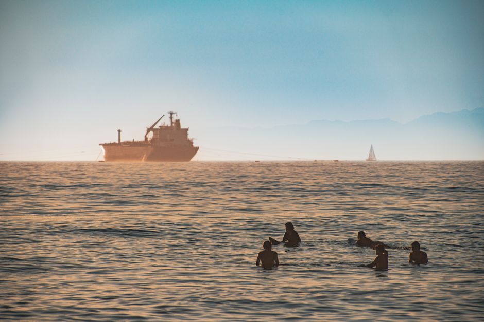 Silhouetted figures in the ocean as a distant ship and sailboat float during a peaceful sunset.