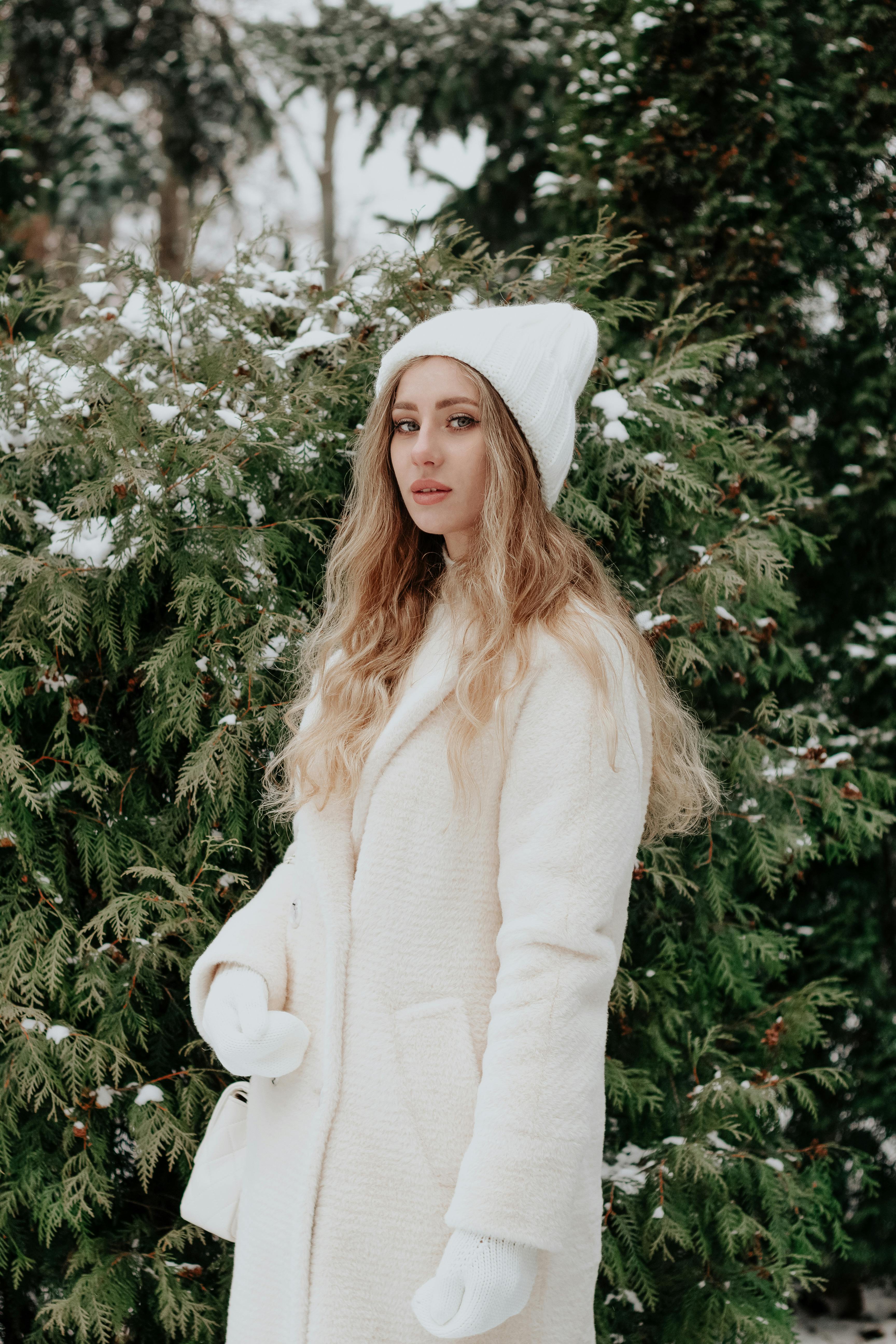 Stylish woman in winter coat and hat poses outdoors amid snowy trees.