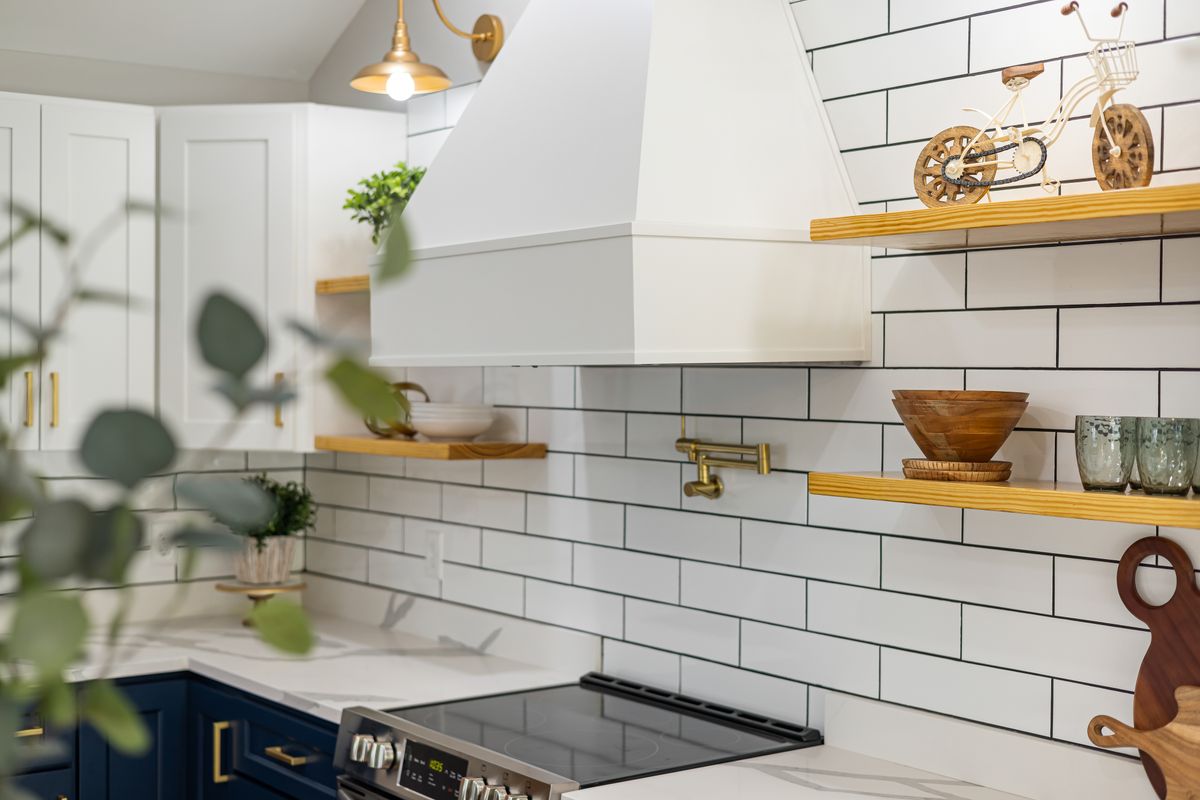 A white tiled kitchen with brass fixtures, open wood shelves and navy cabinets