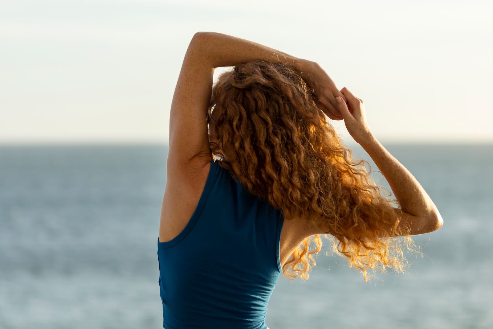 Woman with long curly red hair in a teal tank top stretching her arms overhead beside the ocean in warm sunlight