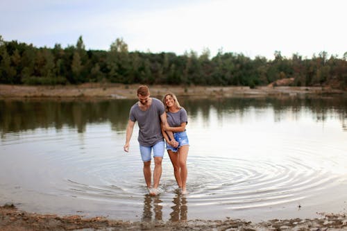 Pasangan Mengenakan Kaos Abu Abu Berjalan Di Atas Air Dangkal Dan Tersenyum