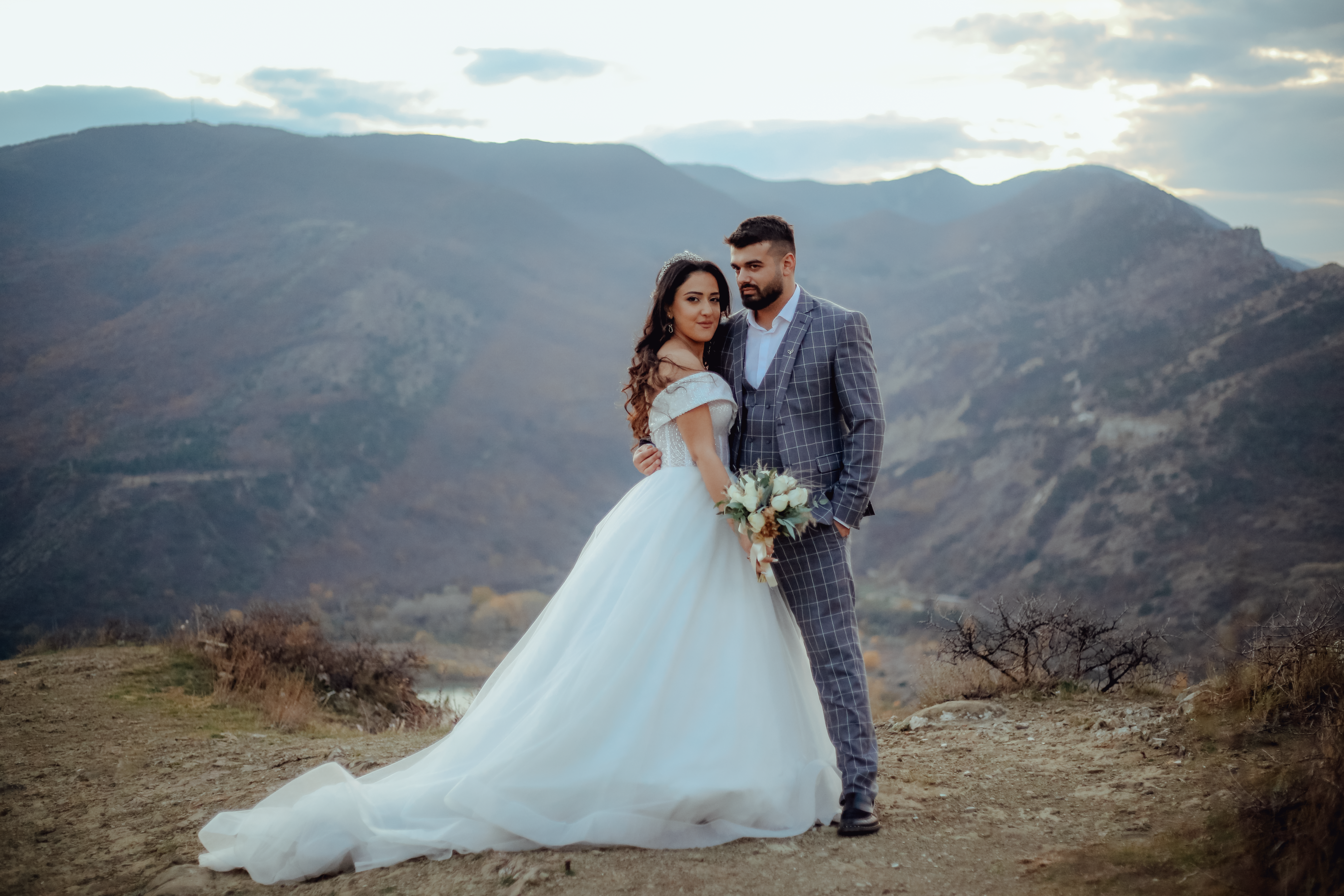 A couple embraces in wedding attire on a mountain top at sunset, symbolizing love and commitment.