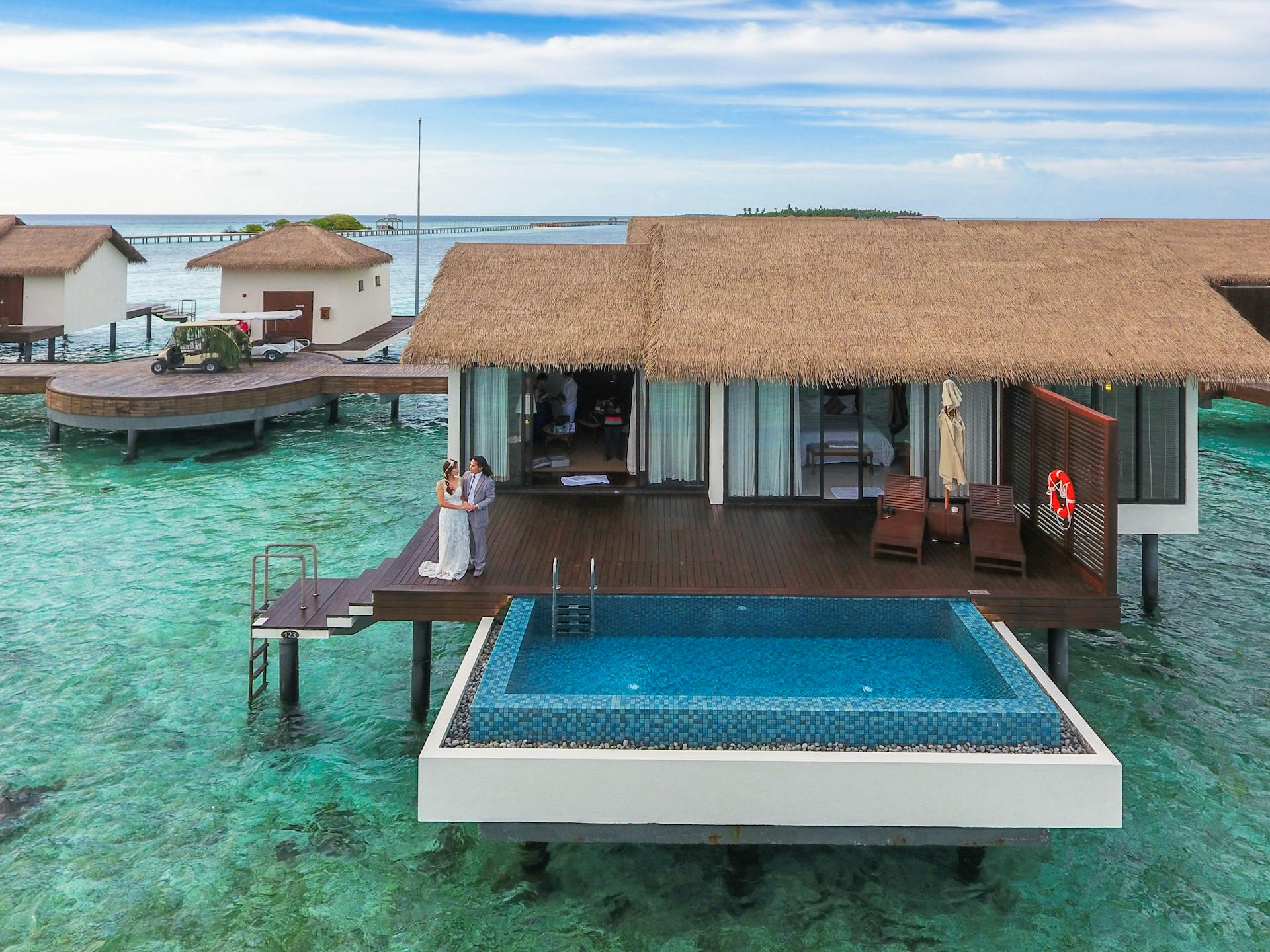 A romantic couple stands on a luxury bungalow deck with an infinity pool over the turquoise ocean.