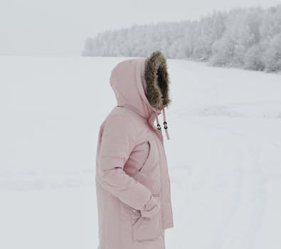 A woman wearing a pink coat stands in a serene snowy landscape, embracing winter's chill.