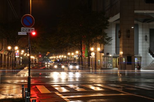 A bustling urban street scene at night with cars, streetlights, and wet roads.
