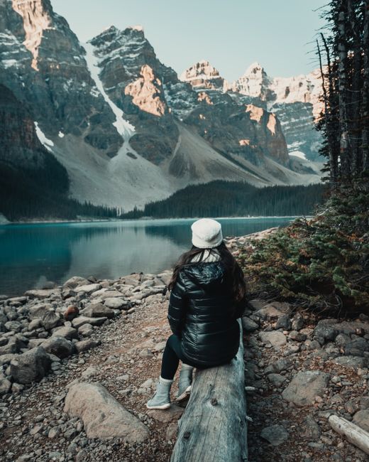 Woman sitting on a log by Moraine Lake, surrounded by snow-capped mountains and pine trees.