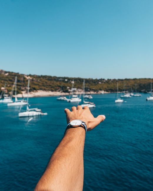 A hand reaching out towards sailboats on a crystal clear sea under a bright blue sky.