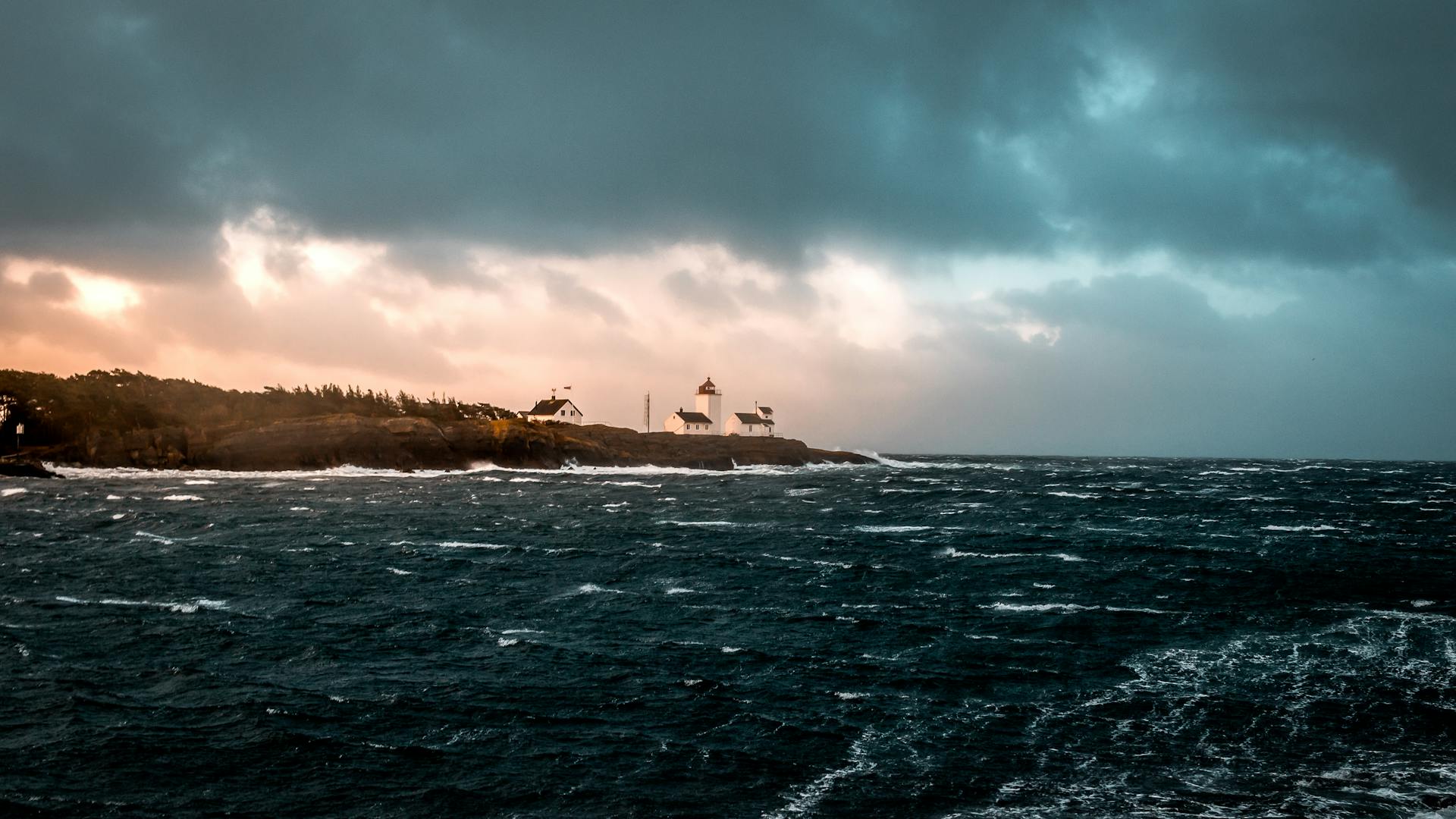 Stunning seascape with a lighthouse on a rocky island during sunset. Waves crash against the shore under a dramatic sky.