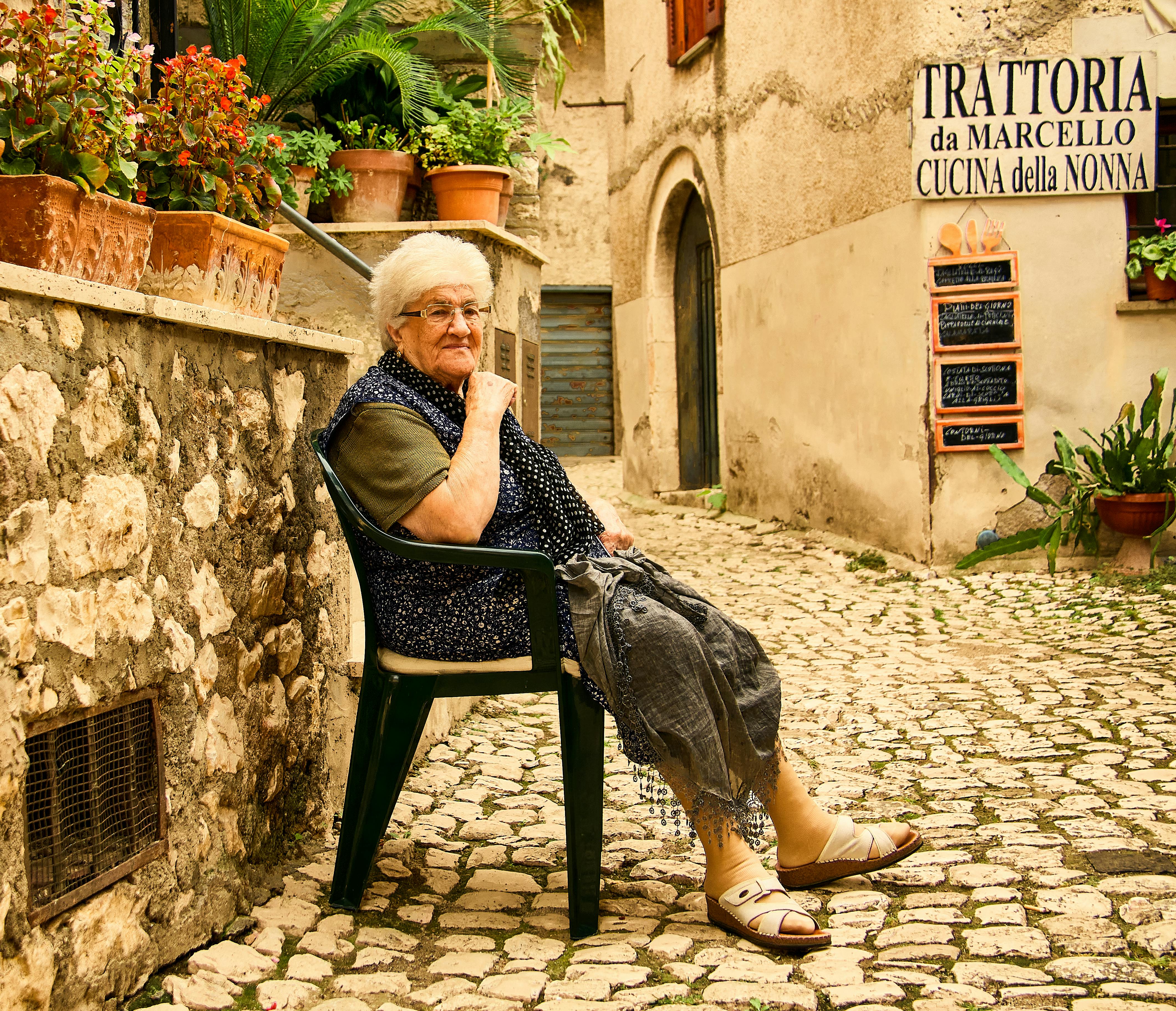 Senior woman in Italian street, sitting by trattoria, surrounded by potted plants.