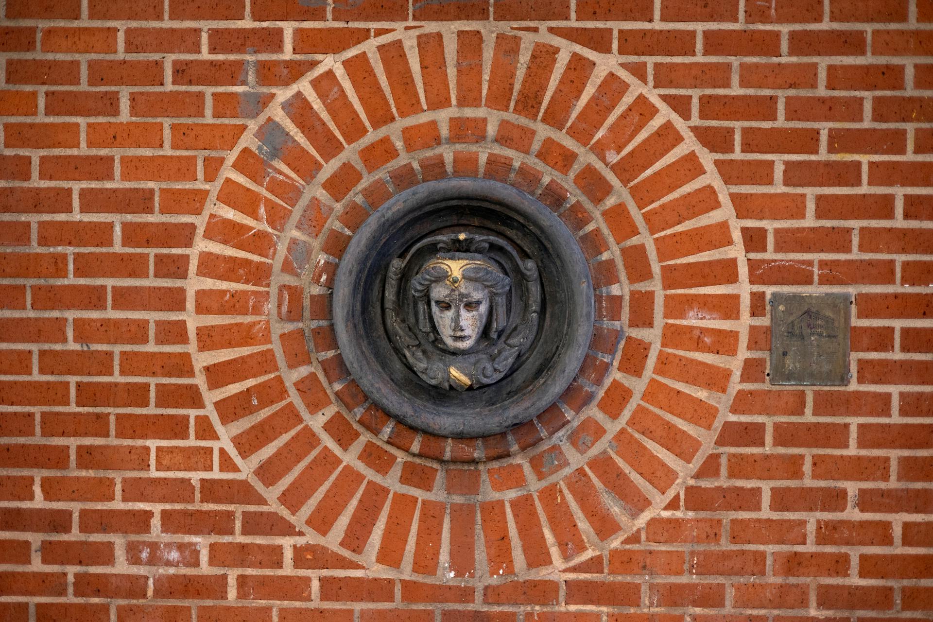 A detailed decorative stone face medallion embedded in a red brick wall, showcasing artistic design.