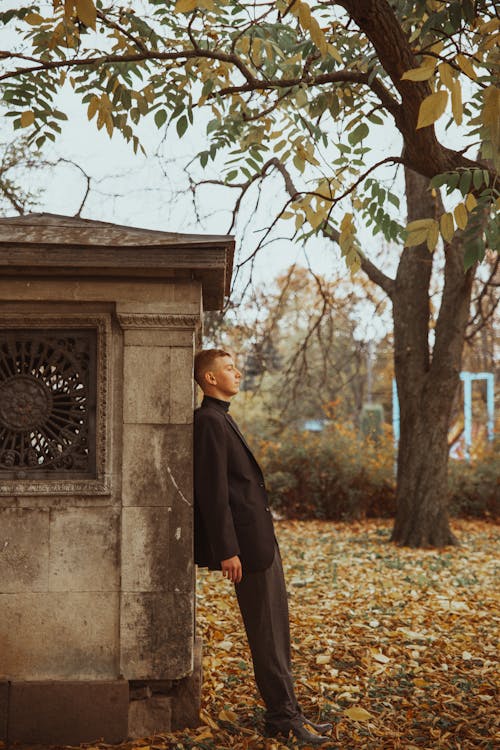 Elegant Young Man Leaning against a Building next to a Tree in Autumn 