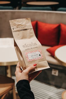 Hand holding a Skola coffee bag inside a cozy cafe with red cushions.