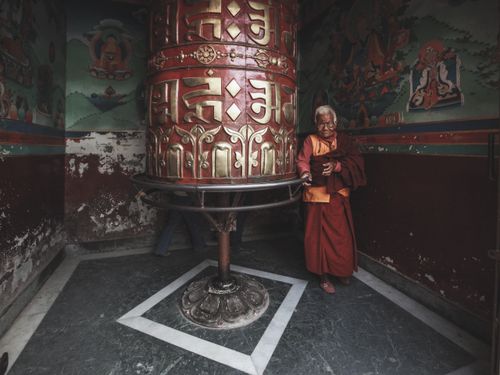 Free Elderly monk beside a large prayer wheel inside a richly decorated monastery room. Stock Photo