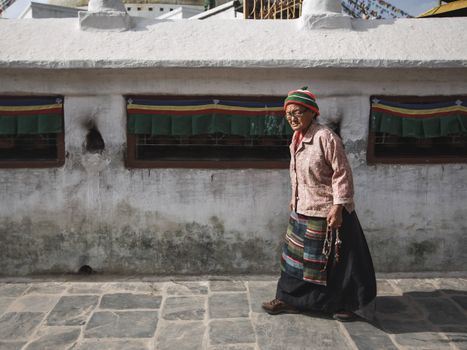 Senior woman in traditional clothing walks beside stone structure outdoors.