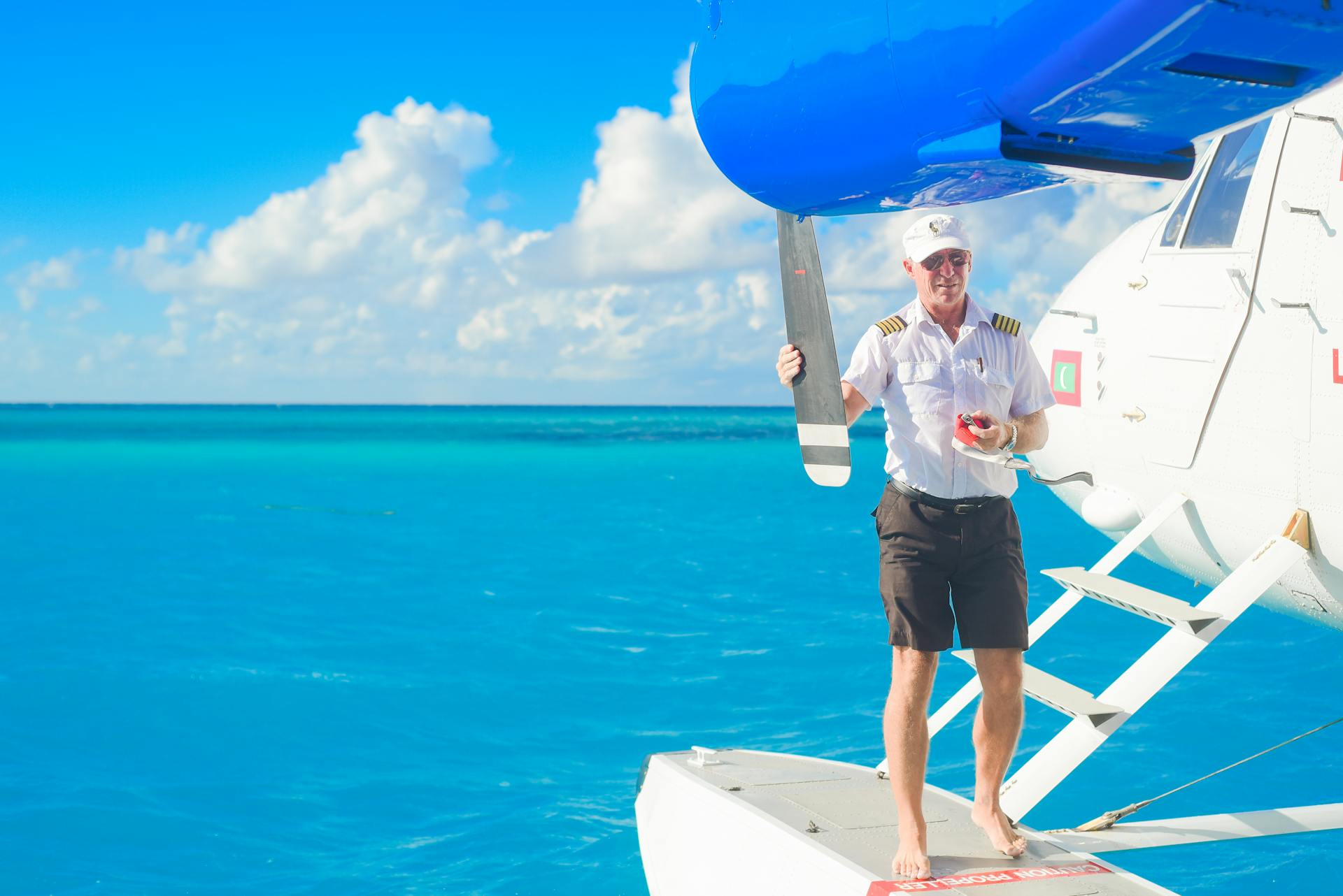 A pilot standing on a seaplane in the clear blue ocean with a bright sky and clouds.